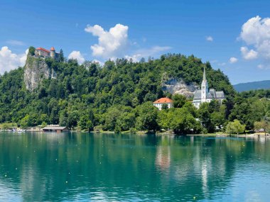 Bled Gölü, Slovenya - 28 Haziran 2024: St. Martin Kilisesi 'nin gri-beyaz mozaik çatısı mavi bulutların altında yeşil yapraklarla çevrili. Beyaz kayalıklar arkada, kalenin üstünde.