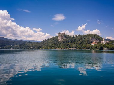 Bled Gölü, Slovenya - 28 Haziran 2024: Gölün suları üzerinde geniş bir manzara ortasındaki kayanın üzerinde kale ve sağdaki St. Martin Kilisesi mavi bulutların altında yansıtılmış.