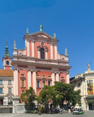 Ljubljana, Slovenya - 28 Haziran 2024: Kırmızı ve pembe tarihi Fransisken Kilisesi, Anma Anıtı anıtsal ön cephesi, heykel Preseren Meydanı 'nda mavi gökyüzü altında. Yeşil yapraklar ve insanlar