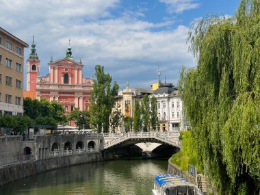 Ljubljana, Slovenya - 28 Haziran 2024: Kırmızı ve pembe tarihi Fransisken kilisesi heykel ile birlikte anıtsal ön cephe. Ljubljanica Nehri 'nin karşısından turne teknesi ve yeşil yapraklarla görüldü.