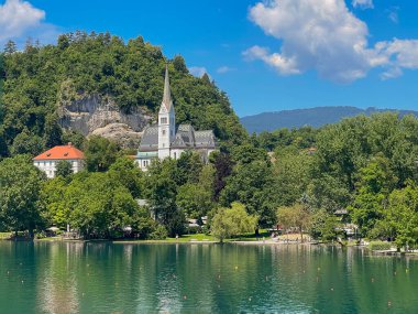 Bled Gölü, Slovenya - 28 Haziran 2024: St. Martin Kilisesi 'nin gri-beyaz mozaik çatısı mavi bulutların altında yeşil yapraklarla çevrili. Beyaz kayalıklar arkada. Geniş beyaz bölge ikametgahı ve yan taraftaki ofis.