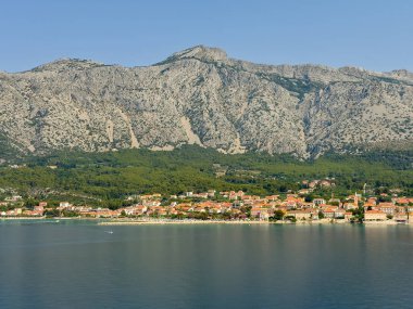 Orebic, Croatia - June 30, 2024: Yacht harbor behind pier and cityscape to the right. Gray rocky mountain range in the back with forested foot. Long sandy Adriatic coastline with hotels clipart