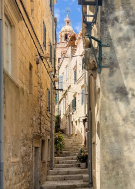 Korcula, Hırvatistan - 30 Haziran 2024: Surlardan batıya, Kalafata ara sokağına baktığımızda, bu dar sarı taştan kanyon boyunca yükselen merdivenleri görüyoruz. Aziz Marks Katedral Kulesi 'nin üzerinde mavi gökyüzüne bakan kubbe var.