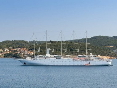 Korcula, Hırvatistan - 30 Haziran 2024: Beyaz Rüzgar Sörfü yolcu gemisi, beş direği ile kasabanın önüne demirledi. Ada kıyısındaki yeşil orman tepeleri ve sahile yakın binalar.