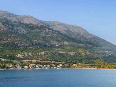 Korcula, Hırvatistan - 30 Haziran 2024: Kuciste 'nin yukarısındaki ana kıyı şeridi ve arkasında yüksek ormanlık bir dağ var. Sahil boyunca birkaç bina var.