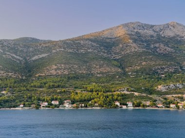 Korcula, Hırvatistan - 30 Haziran 2024: Kuciste 'nin yukarısındaki ana kıyı şeridi ve arkasında yüksek ormanlık bir dağ var. Sahil boyunca birkaç bina var.