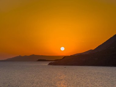 Korcula, Hırvatistan - 30 Haziran 2024: Şehrin batısından gün batımı görüldü. Turuncu gökyüzünde, orman adalarının koyu yamaçlarında hafif ateş topu belirlendi.