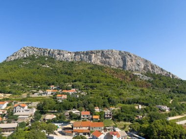 Bölünme, Hırvatistan - 1 Temmuz 2024: Kuzey Klis Kalesi, yeşil ormanların üzerinde gri dağ tepeleri ve kırmızı çatıları olan beyaz evlerin üzerinde yer alıyor