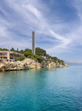 Split, Hırvatistan - 1 Temmuz 2024: Uzun Sailor Deniz Feneri ve limanın doğu yakasında feribot terminalinin arkasında mavi bulutların altında iletişim kulesi. Rocky kıyı şeridi, Kavala plajı ve uzun yeşil ağaçlar.