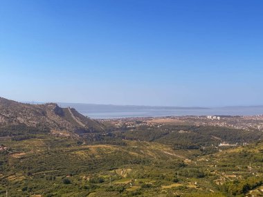 Split, Hırvatistan - 1 Temmuz 2024: Klis Kalesi 'nden Adriyatik Denizi' nin mavi gökyüzü altında yer aldığı yeşil orman ve tarım sahaları manzarası üzerinde görülen şehir manzarası. Doğuda bir dağ kanadı var.