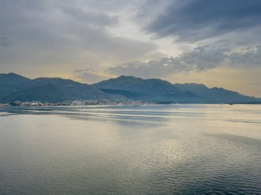 Kotor Körfezi, Karadağ - 2 Temmuz 2024: Aşağı körfezin doğu ucundaki Tivat kasabasında gün doğumu gri bulutların altında sarı bir gökyüzünü gösteriyor. Karanlık dağlar ve şehir manzarası