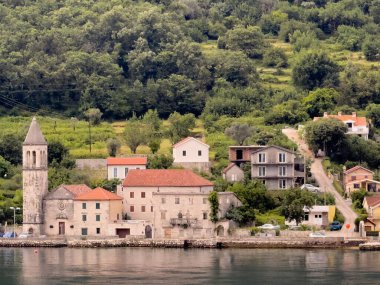 Kotor Körfezi, Karadağ - 2 Temmuz 2024: Donji Stoliv 'deki Saint Basil Kilisesi kıyı şeridinde ve sarp dağ yamacının önündeki kırmızı çatılı evlerle çevrili