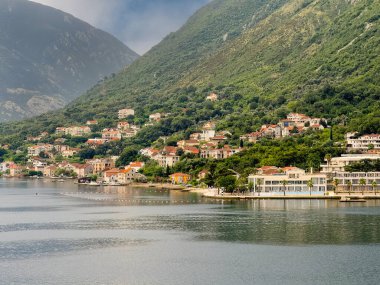 Bay of Kotor, Montenegro - July 2, 2024: Hyatt Regency Hotel, NE corner, at Donji Stoliv built on peninsula coastline in cityscape of red-roofed buildings up the green mountain. Beach and boats clipart