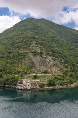 Bay of Kotor, Montenegro - July 2, 2024: At the north exit of the narrow channel on the east shoreline, we find the Church of Our Lady of Angels Unesco-protected building with a light tower beacon. Green mountain slopes clipart