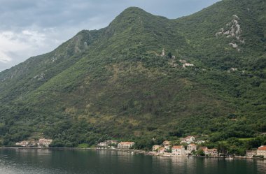 Kotor Körfezi, Karadağ - 2 Temmuz 2024: Körfezin kuzey kıyısında Donji Stoliv cemaati. Su rıhtımına yakın küçük tekneleri olan bej kırmızı çatılı evler. Ormanlı dağ yamacı