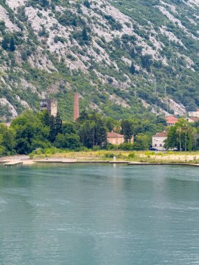 Kotor, Karadağ - 2 Temmuz 2024: Small Beach 'in arkasındaki terk edilmiş AR Industrial binaları ve yeni konut evleri. Arkalarındaki Lovcen Dağı 'ndan gri kaya duvarı. Körfez suyu önde.