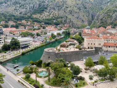 Kotor, Montenegro - July 2, 2024: Saint Nicholas church domes over other red-roofed buildings of historic downtown. Ramparts upfront and fountain. Gray rock mountain range in back clipart