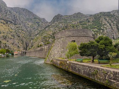 Kotor, Karadağ - 2 Temmuz 2024: NW gözcü köşeli tarihi şehir merkezi surları. Öndeki yeşil park, arkadaki dağ sırası mavi bulutların altında.