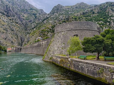 Kotor, Karadağ - 2 Temmuz 2024: NW gözcü köşeli tarihi şehir merkezi surları. Öndeki yeşil park, arkadaki dağ sırası mavi bulutların altında.