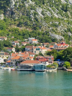 Kotor, Karadağ - 2 Temmuz 2024: Kırmızı çatılı binası olan ve körfezin güney kıyısında arkasında gri ormanlık kayalık dağ bulunan Galion Restoranı