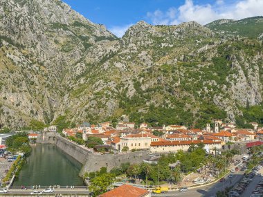 Kotor, Karadağ - 2 Temmuz 2024: NE köşesi ve kuzey surları tarihi şehir merkezinde gri kayalık dağ kanatları ile mavi bulutların altında. Kırmızı çatılı binalar. Kilise ve kale
