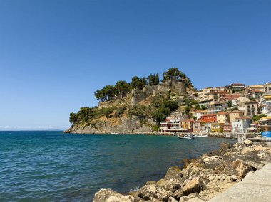 Parga, Yunanistan - 3 Temmuz 2024: Venedik şatosu ve onun yeşil yaprakları mavi gökyüzü altında İyon Denizi üzerindeki kayalarda. Yeşilimsi su ve çok katlı evler ve otel binaları duvarı. Sudaki küçük motorlu tekneler