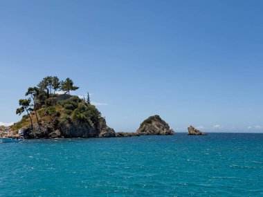 Parga, Yunanistan - 3 Temmuz 2024: Saint Prokopios Kutsal Şapel adası, mavi gökyüzünün altında sadece birkaç metre ötede bulunan uzun yeşil yapraklı ağaçlarıyla. Güney noktasındaki kayalar Azure deniz suyunda