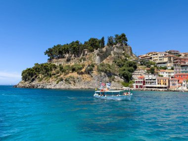 Parga, Yunanistan - 3 Temmuz 2024: Venedik şatosu ve onun yeşil yaprakları mavi gökyüzü altında İyon Denizi üzerindeki kayalarda. Gök mavisi deniz suyunda bir turist teknesi. Çok katlı evler ve otel binaları..