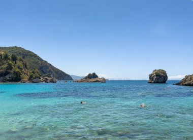 Parga, Yunanistan - 3 Temmuz 2024: İyon Denizi 'nde yüzücüler mavi gökyüzünün altındaki mavi deniz suyundan çıkan kayalarla