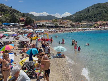 Parga, Yunanistan - 3 Temmuz 2024: Mavi gökyüzünün altında ve dışında insanlarla dolu bir plaj. İyon Denizi 'ne bir sürü şemsiye ve orman tepeleri iniyor. Bistrolar duvarı ve daha fazlası sahil yolunda.