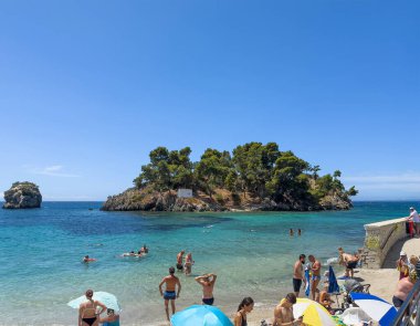 Parga, Yunanistan - 3 Temmuz 2024: Mavi gökyüzünün altında ve dışında insanlarla dolu bir plaj. Aziz Prokopios Kutsal Şapeli mavi gökyüzünün altında, adasında.
