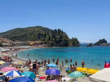 Parga, Yunanistan - 3 Temmuz 2024: Mavi gökyüzünün altında ve dışında insanlarla dolu bir plaj. İyon Denizi 'ne bir sürü şemsiye ve orman tepeleri iniyor. Bistrolar duvarı ve daha fazlası sahil yolunda.