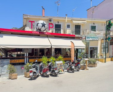 Parga, Yunanistan - 3 Temmuz 2024: St. Nicholas Kilisesi 'nin karşısındaki Cafe Grill Top, komşusu bir eczane. Sembol olarak büyük şişme futbol topu. İş yerinin önünde motosikletler