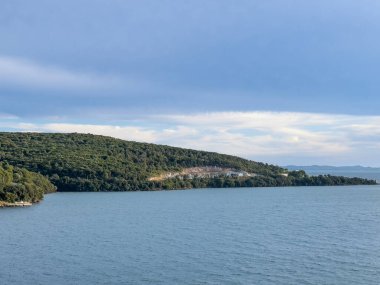 Igoumenitsa, Yunanistan - 3 Temmuz 2024: Limanın dışındaki güney yarımadasının tepesindeki taş ocağı. Mavi bulutların altında Ormanlı tepe ile yeşil manzara