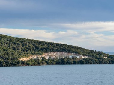 Igoumenitsa, Yunanistan - 3 Temmuz 2024: Limanın dışındaki güney yarımadasının tepesindeki taş ocağı. Mavi bulutların altında Ormanlı tepe ile yeşil manzara