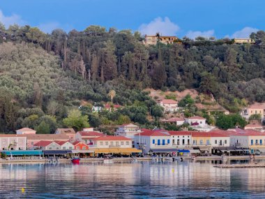 Katakolo, Greece - July 4, 2024: Boardwalk along the harbor show line of commercial buildings with forest on hill with sprinkled mansions in back. Small boats along the quay.  clipart