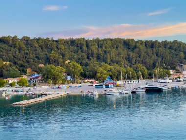 Katakolo, Yunanistan - 4 Temmuz 2024: Katakolo Yacht Marina, Ilia, rıhtım boyunca uzanan ticari binaların arkasındaki ormanlık kıyı şeridi ile mavi sabah gökyüzü altında turuncu gün ışığı bulutu ile renklendirilmiş
