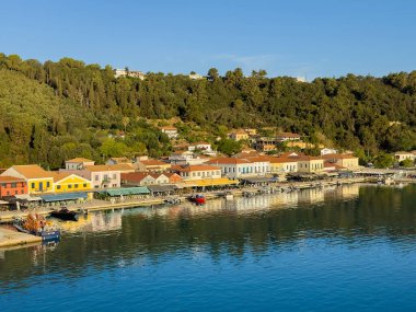 Katakolo, Yunanistan - 4 Temmuz 2024: Yat limanındaki Boardwalk 'ta çoğunlukla perakende, restoran ve bistrolar olmak üzere ticari binalar sergileniyor. Ormandaki tepe mavi gökyüzünün altında. Rıhtımda küçük tekneler