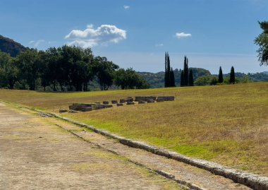 Olympia, Yunanistan - 4 Temmuz 2024: Olimpiyat Oyunlarının doğduğu yer. Mavi bulutlu bir manzaranın altında küçük bir binanın bulunduğu eğimli tarafı olan antik bir stadyum spor alanıydı. Arkasında yeşil ağaçlar