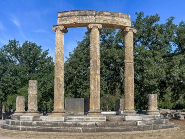 Olympia, Yunanistan - 4 Temmuz 2024: Olimpiyat Oyunlarının doğduğu yer. Mavi gökyüzünün altındaki yeşil ağaçlarla desteklenen Filipinli harabeleri.