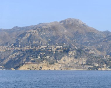 Taormina, Sicilya, İtalya - 5 Temmuz 2024: Isola Bella kayalıklarından Mendolia sahiline, Mazzaro kayalıklarına kadar uzanan Taormina kıyıları. Mavi gökyüzünün altındaki dağ sırasının kenarındaki şehir manzarası