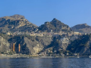 Taormina, Sicilya, İtalya - 5 Temmuz 2024: Kayalık kıyı şeridi üzerindeki Taormina merkezi şehir merkezi ve Lorenzina plajı iki dev deniz trafiği turuncu feneriyle. Mavi gökyüzünün altındaki yüksek dağlarda daha fazla bina