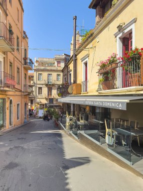Taormina, Sicilya, İtalya - 5 Temmuz 2024: Dar sokak manzaralı. Bistro Santa Domenica önü, sandalyeler hala masanın üzerinde, balkonda kırmızı çiçekler. Diğer taraftaki General Finansal Sigorta Ofisi. 