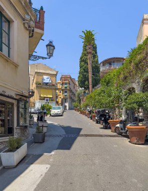 Taormina, Sicilya, İtalya - 5 Temmuz 2024: Sesto Pompeo üzerinden Corso Umberto 'dan park etmiş arabalar ve motosikletlerle görüldü. Bir tarafta yeşil ağaçlar var. Mavi gökyüzünün altında birkaç apartman.