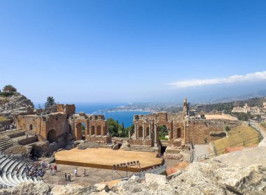 Taormina, Sicilya, İtalya - 5 Temmuz 2024: Antik Yunan tiyatrosu veya Teatro antico. Sahne ve sahne arkası harabeleriyle kuzeyden geniş manzara manzarası, ufukta mavi deniz ve kıyı şeridi. Mevcut insanlar
