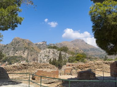 Taormina, Sicilya, İtalya - 5 Temmuz 2024: Antik Yunan tiyatrosunun veya Teatro Antico 'nun SE kalıntıları. Mavi bulutların altında alçak duvarlar, arkada dağ