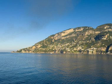 Amalfi, Salerno, İtalya - 6 Temmuz 2024: Yeşil dağ sırası ve mavi gökyüzünün altındaki bej kayalık kayalıklar ile şehrin batı kıyı şeridi. Kanatlara binalar serpilmişti.. 
