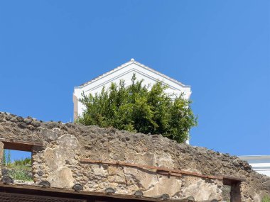Pompeii, Campania, İtalya - 6 Temmuz 2024: Via Dell 'Abbondanza' nın kuzeyindeki Antik Roma şehri kalıntıları. Modern beyaz bina mavi gökyüzünün altındaki yeşil ağaç yapraklarının üzerinde.