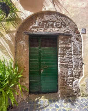 Trevignano Romano, Lazio, İtalya - 7 Temmuz 2024 Via Umberto 54, önünde eğrelti otu saksısı bulunan, taş yay çerçeveli ağır ahşap kapı. Sokak yüzeyi: küçük kaldırım taşları