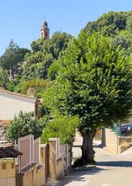 Ocana, Korsika, Fransa - 8 Temmuz 2024: Yerel kilise kulesi yeşil ağaç yapraklarının üzerinde dikiz yapıyor. 